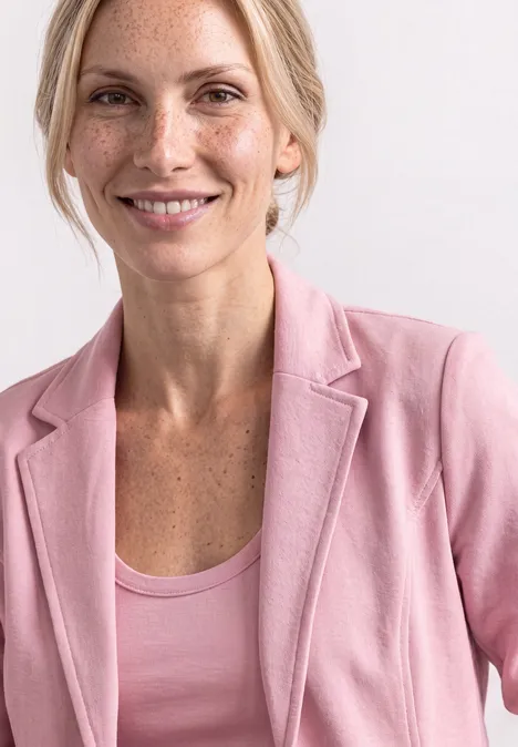 Eine Frau mit Sommersprossen trägt einen rosa Blazer und Top und lächelt vor hellem Hintergrund.