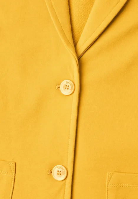 Nahaufnahme eines senfgelben Blazers mit zwei Knöpfen, Reverskragen und Taschen.