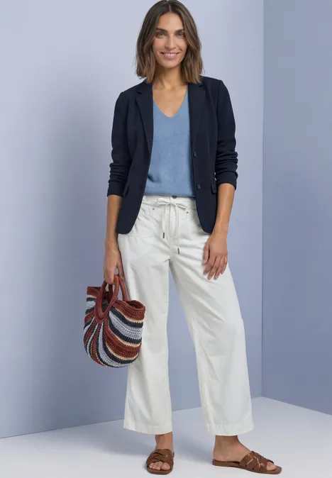 Frau mit dunkelblauem Blazer, hellblauem Top, weißer Culotte, braunen Sandalen und gestreifter Tasche.
