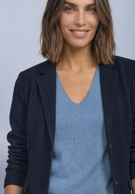 Frau mit brauen, schulterlangen Haaren, blauem V-Ausschnitt-Pullover und dunkelblauem Blazer, leichtes Lächeln.