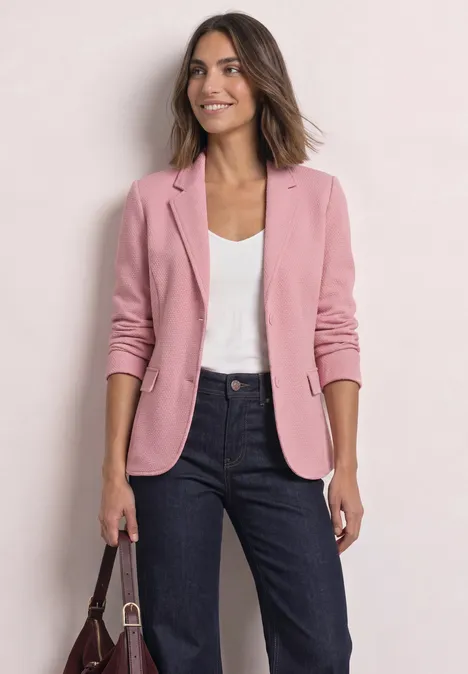 Vrouw in roze blazer, wit shirt en donkere jeans, houdt een handtas voor een lichte achtergrond.
