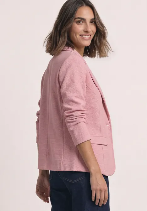 Vrouw in een roze blazer en spijkerbroek lacht schuin naar de camera.