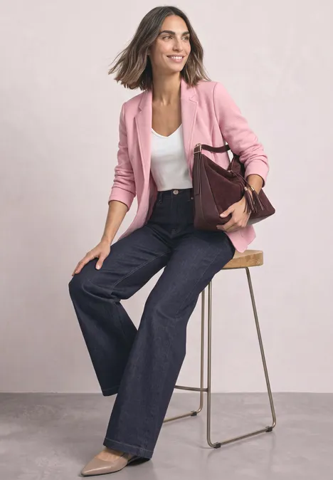 Vrouw in roze blazer, wit topje en wijde jeans zit op een kruk en houdt een bordeaux handtas.