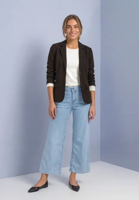 Eine junge Frau in schwarzem Blazer, weißem T-Shirt, weiten Jeans und schwarzen Schuhen vor hellem Hintergrund.