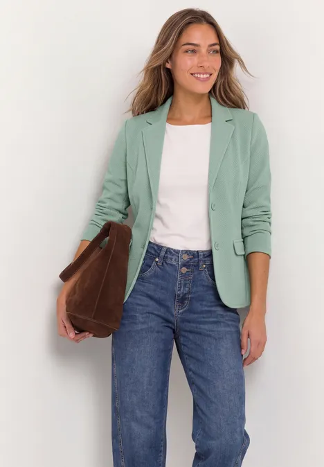 Frau in mintgrünem Blazer, weißem Shirt, Jeans und mit brauner Tasche.