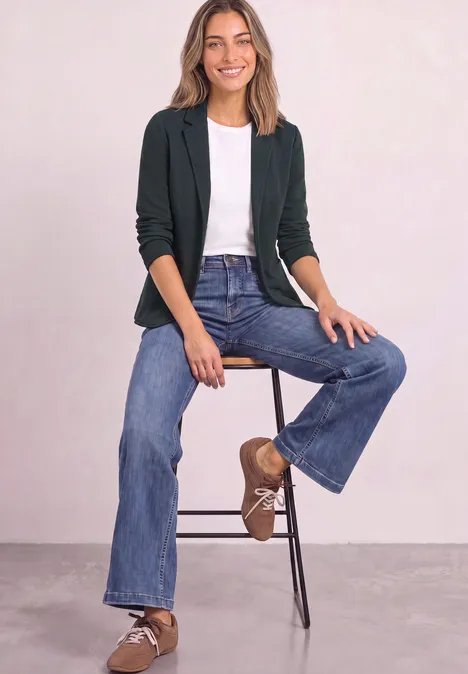 Eine Frau sitzt auf einem Hocker, trägt Blazer, weißes T-Shirt, weite Jeans und braune Schnürschuhe.