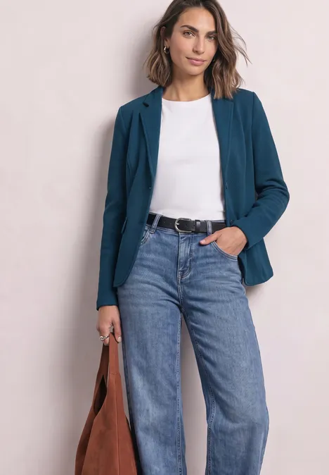 Frau im teal Blazer, weißem Shirt, Jeans und brauner Tasche