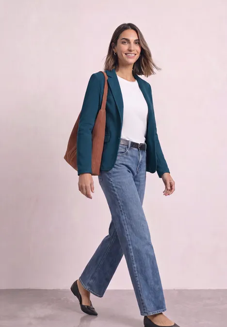 Eine lächelnde Frau im grünen Blazer, weißem Shirt und Jeans trägt eine braune Tasche vor rosa Hintergrund.
