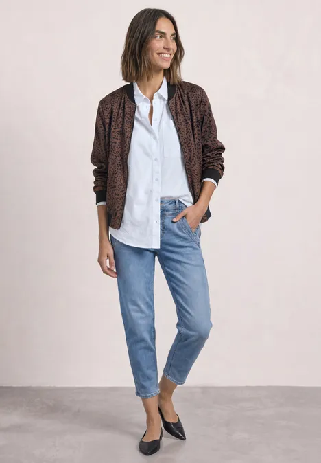 Eine lächelnde Frau in gestreiftem Hemd, gemustertem Blouson, hellen Jeans und schwarzen Ballerinas.