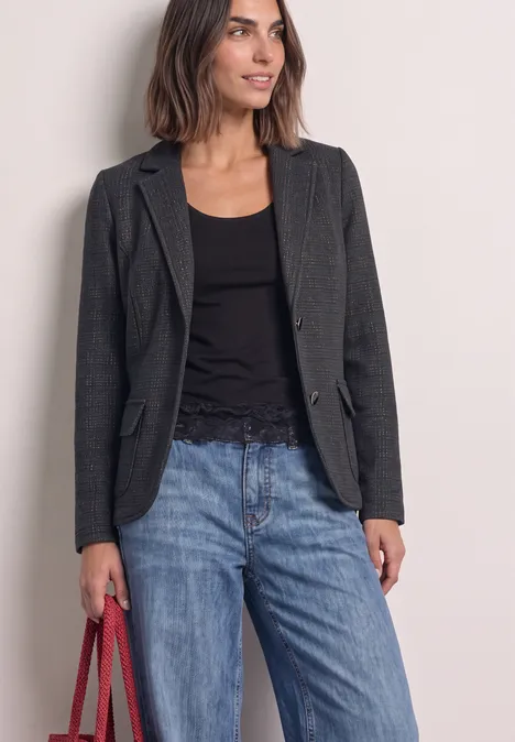 Eine Frau trägt ein dunkles Sakko, ein schwarzes Oberteil, blaue Jeans und hält eine rote Tasche.