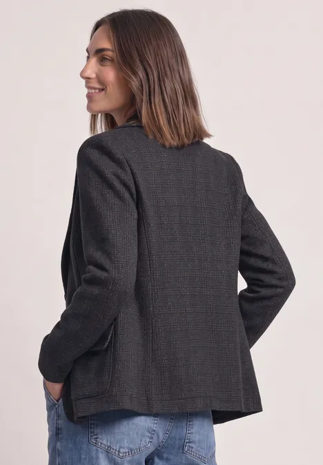 Frau mit schulterlangem Haar in dunkelgrauem Blazer und Jeans, seitlich stehend mit Händen in den Taschen.