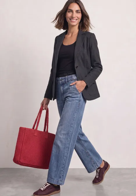 Frau in schwarzem Blazer und Jeans trägt rote Tasche und geht lächelnd.