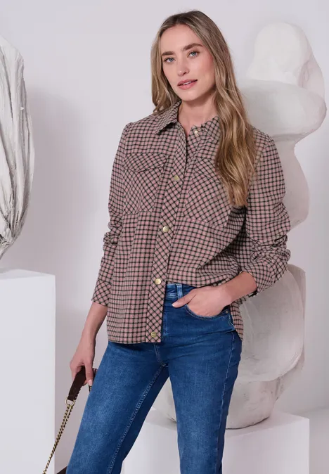 Junge Frau trägt karierte Jacke und Jeans, steht vor einer Skulptur, lächelt und hält eine Tasche.