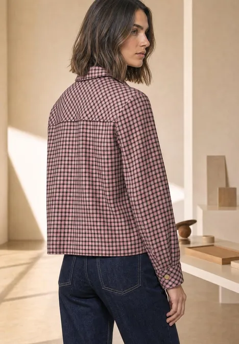 Frau in karierter Jacke und Jeans, steht seitlich in hellem Raum.