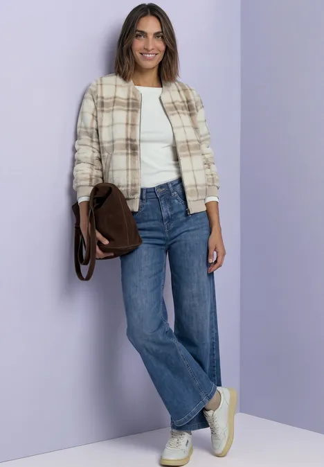 Eine Frau in Jeans, weißem Top und kariertem Jackett steht lässig mit brauner Tasche.