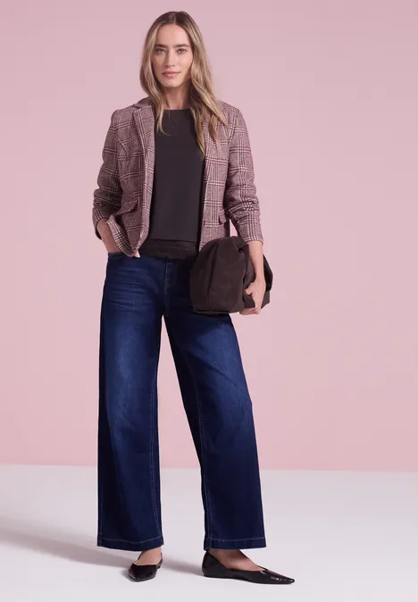 Een vrouw draagt een blazer, zwart shirt, wijd jeans en houdt een tas vast voor een roze achtergrond.