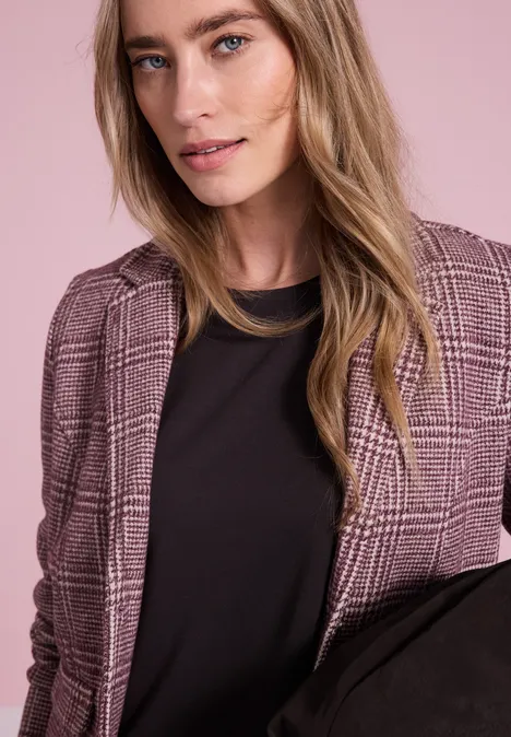 Jonge vrouw met blond haar draagt een zwart t-shirt en een roze geruit colbert voor een pastelkleurige achtergrond.