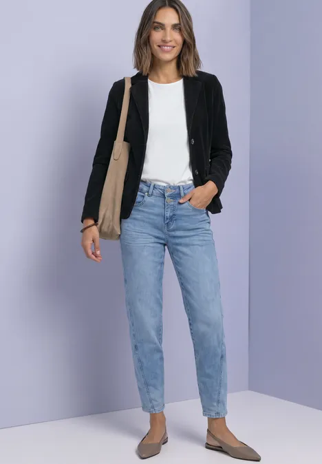 Femme devant un fond lila-gris, en veste noire, t-shirt blanc, jean bleu clair, ballerines et sac beige.