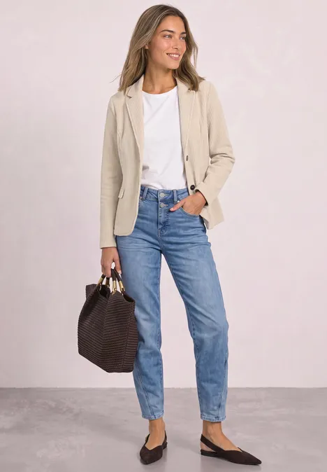 Femme en blazer beige, t-shirt blanc, jean bleu et ballerines noires, portant un sac à main foncé.