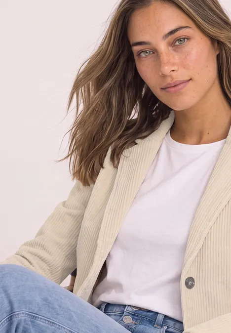 Femme aux cheveux bruns en t-shirt blanc et blazer beige devant un fond clair.