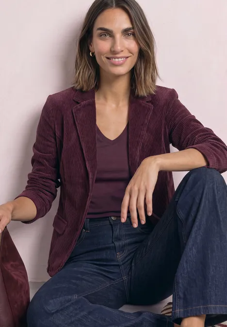 Femme assise souriante, blazer bordeaux, top sombre et jean large, pose décontractée sur fond clair.