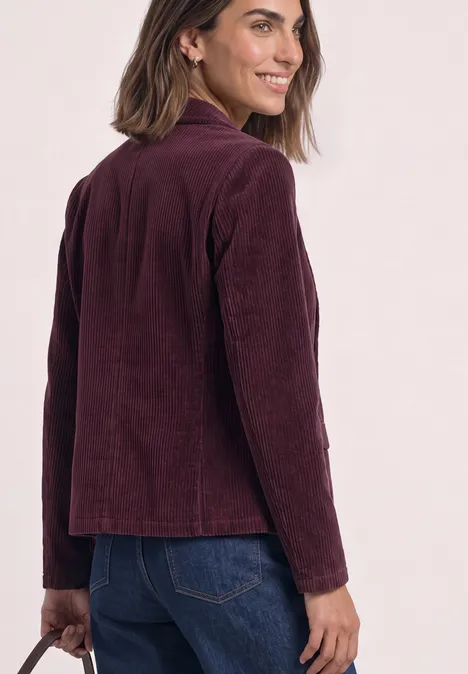 Femme portant une veste en velours côtelé bordeaux et un jean, souriante en regardant par-dessus l'épaule.