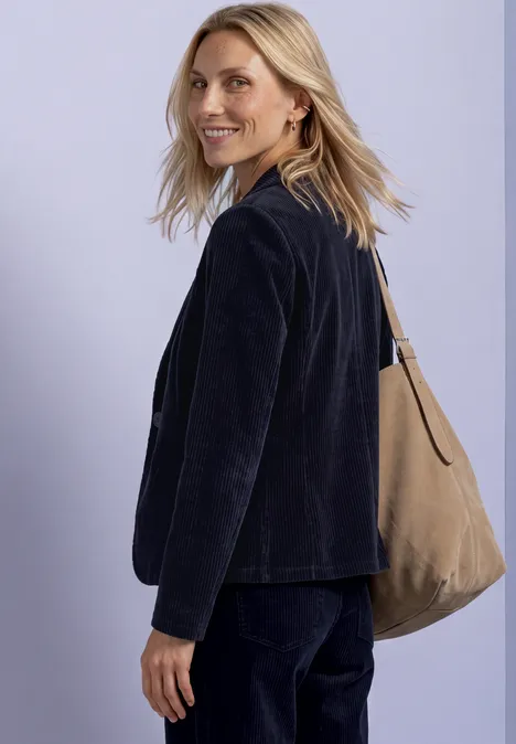 Une femme blonde souriante en tenue sombre avec un sac beige à l’épaule devant un fond clair.