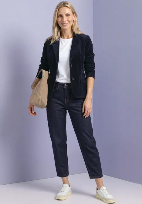 Une femme en veste bleu marine, t-shirt blanc, jean et baskets blanches, sac beige, devant un fond violet clair.