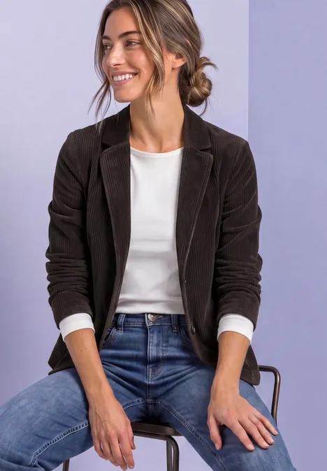 Femme assise souriante sur une chaise, portant un blazer noir, un t-shirt blanc et un jean bleu sur fond clair.