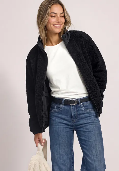 Une femme porte une veste en polaire noire, un t-shirt blanc et un jean, elle sourit en tenant un tissu.