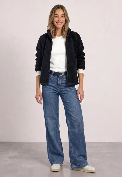 Une femme souriante porte une veste polaire noire, un haut blanc, un jean évasé et des baskets blanches devant un fond gris clair.