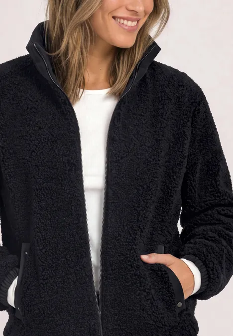 Une femme sourit en portant une veste polaire teddy noire zippée avec poches.