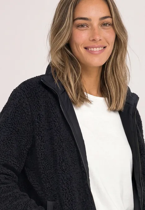 Une femme souriante porte une veste polaire noire et un t-shirt blanc sur fond clair.
