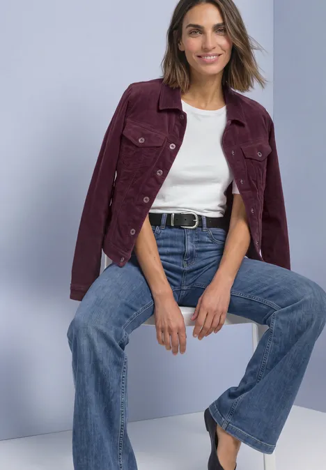 Vrouw zit ontspannen in wijde jeans, wit shirt en bordeaux jas op een lichtgrijze achtergrond.