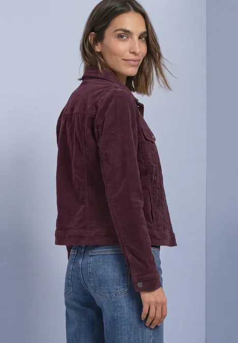 Vrouw in wijnrode corduroy jas en blauwe jeans, zijwaarts, kijkt naar de camera tegen effen achtergrond.