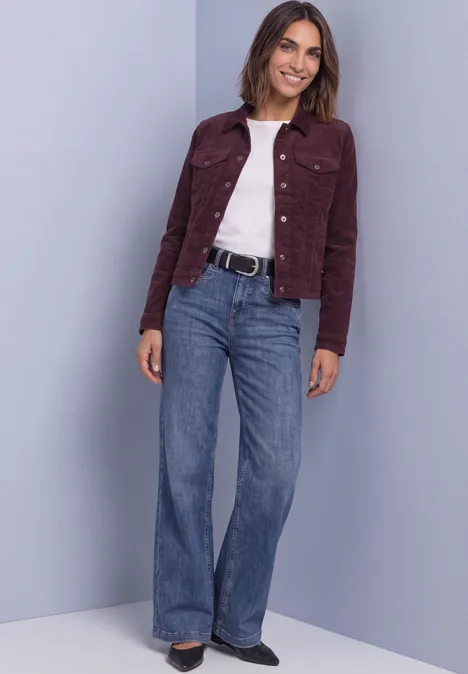 Vrouw in bordeaux corduroyjasje, wit shirt, wijdvallende jeans en zwarte riem voor een neutrale achtergrond.
