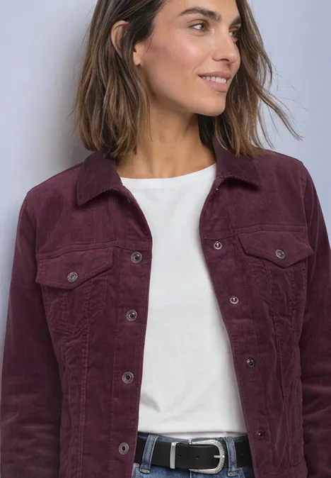 Vrouw in zijaanzicht draagt wit shirt onder paarse corduroy jas met zakken en zwarte riem, glimlacht.