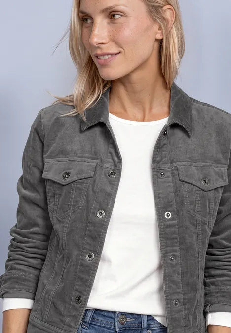 Een vrouw met lichtblond haar draagt een wit T-shirt en een grijze jas.