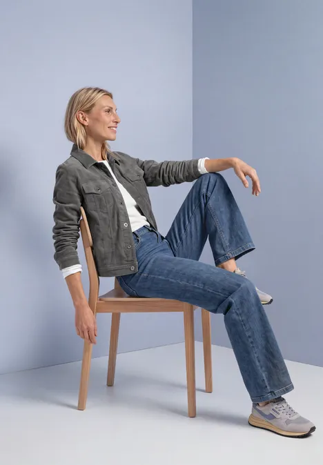 Een blonde vrouw zit ontspannen op een kruk, draagt jeans, witte sneakers en jas tegen een lichtblauwe achtergrond.
