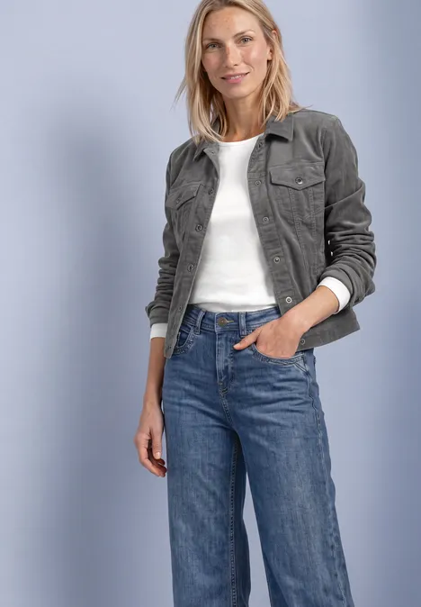 Vrouw in donkergroene corduroy jas, wit topje en blauwe jeans, glimlachend met een hand in de zak.