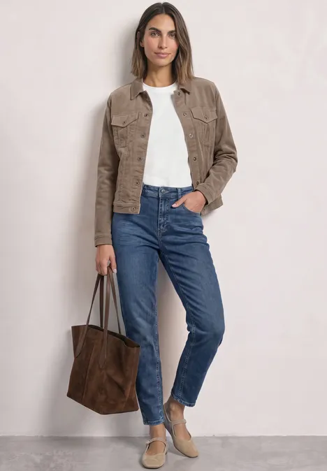 Vrouw in jeans, wit shirt en bruine jas draagt een grote bruine tas voor een neutrale achtergrond.