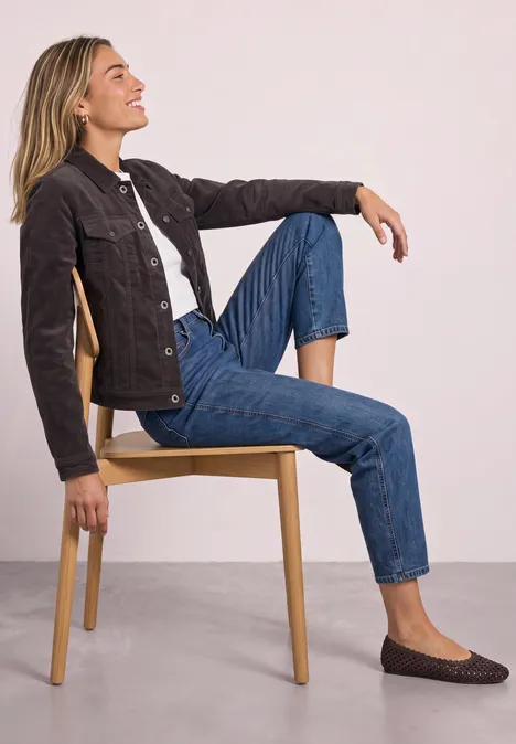 Vrouw zit relaxt op een stoel, leunt achterover, één knie opgetrokken, draagt jeans, jack en platte schoenen.
