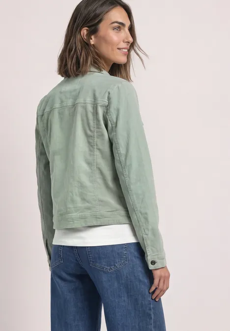 Vrouw van achteren met groene jas en spijkerbroek, kijkt over haar schouder, neutrale achtergrond.