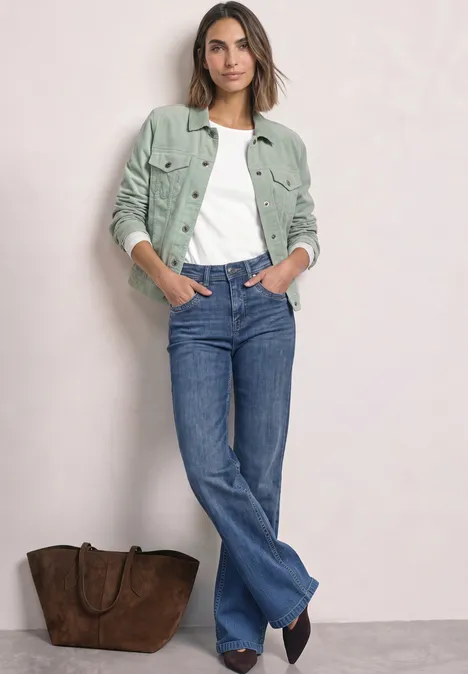 Vrouw met groene jas, wit shirt en blauwe jeans staat voor een lichte achtergrond naast een bruine tas.