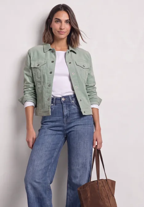 Vrouw in groene corduroy jas, witte top en spijkerbroek met een bruine tas.