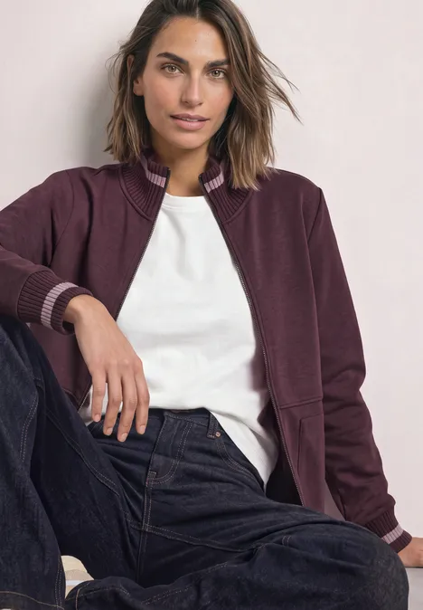 Frau mit kastanienfarbenem Blouson, weißem T-Shirt und Jeans sitzt lässig und blickt selbstbewusst.