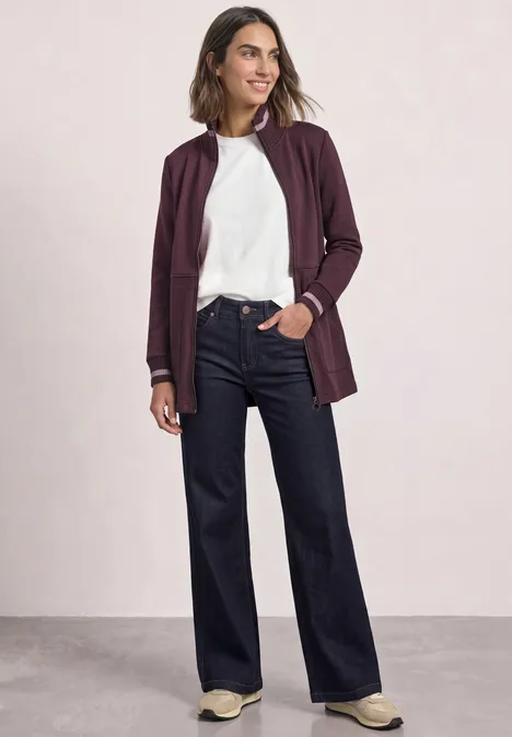 Frau in weißem Shirt, bordeauxfarbener Jacke und weiten, dunklen Jeans, sportliche Sneaker.