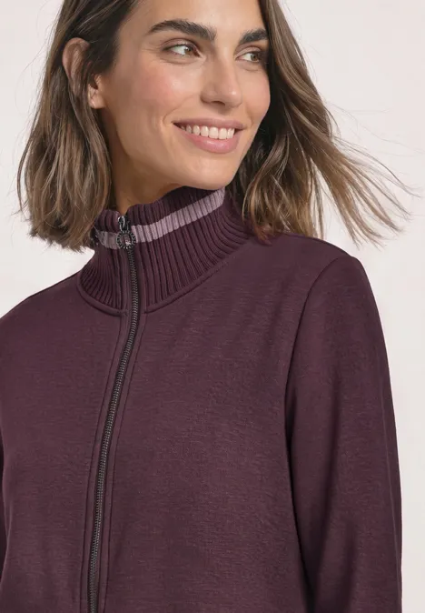 Frau mit schulterlangem Haar trägt einen violetten Pullover mit Reißverschluss und hohem Kragen.