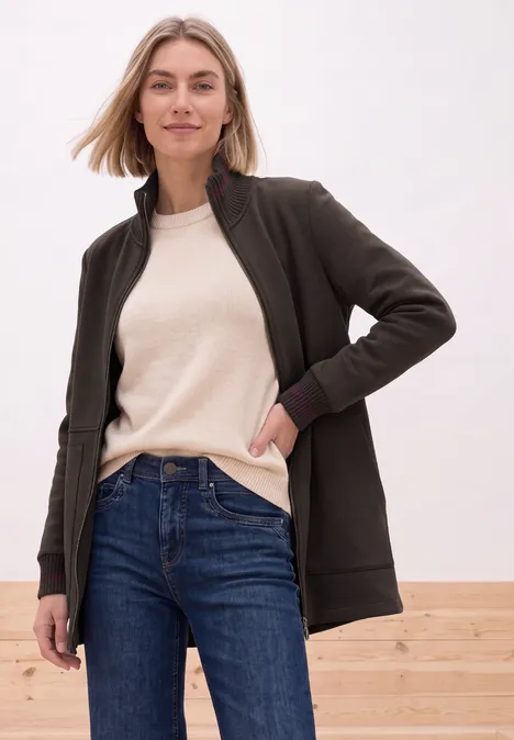 Femme en gilet noir, t-shirt crème et jean, posant de manière détendue devant un mur clair.