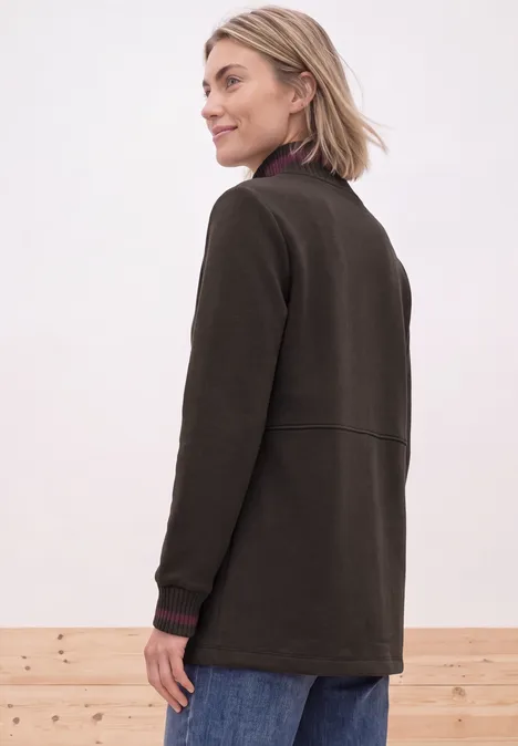 Femme en veste brune vue de profil devant un mur clair.